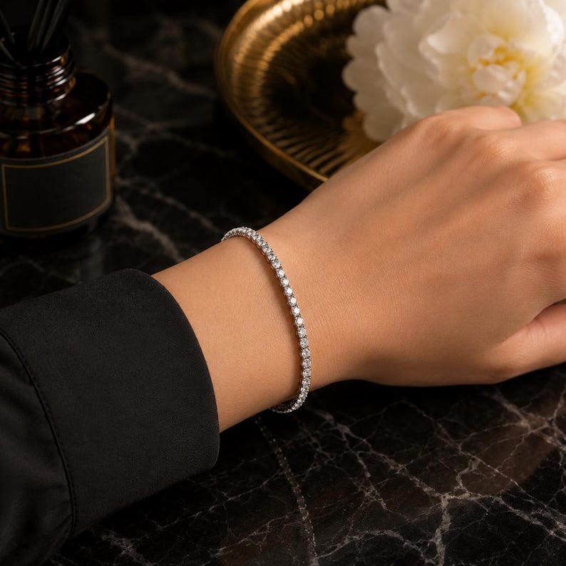 May include: A silver-toned tennis bracelet featuring a continuous row of small, round, clear faceted stones. The bracelet is worn on a wrist, showcasing a flexible metal chain design with a secure clasp mechanism and a polished, reflective finish.