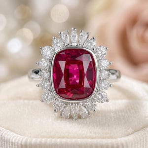 Puede incluir: Un anillo de tono plateado con una gran piedra preciosa de color rojo intenso, de corte cojín rectangular, en el centro. La piedra central está rodeada por un halo de pequeñas piedras blancas claras, redondas y de corte marquesa.