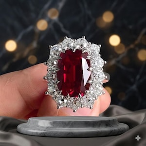 Puede incluir: Un anillo con una gran piedra preciosa rectangular de color rojo intenso en el centro, rodeada por un halo de numerosas piedras blancas pequeñas, redondas y claras engastadas en una banda metálica de aspecto brillante y facetado.