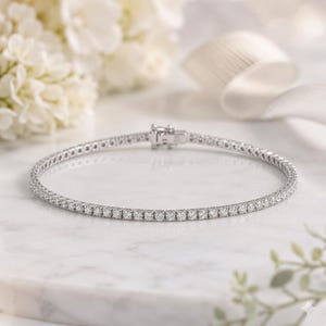 May include: A silver-toned tennis bracelet featuring a continuous row of small, round, clear stones set in a metallic frame. The bracelet includes a secure box clasp mechanism at the top, showcasing a uniform design throughout the entire piece.