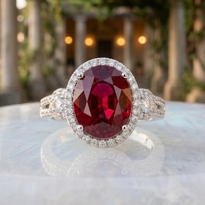 May include: A silver-toned ring featuring a large, oval-cut deep red gemstone at the center. The central stone is surrounded by a halo of small, clear, round-cut stones. The band is also encrusted with small, clear, round-cut stones along the sides.