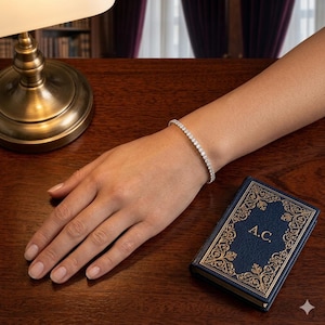 May include: A silver-toned tennis bracelet featuring a continuous row of small, clear, round-cut stones. The bracelet is worn on a wrist, positioned near a small, rectangular, dark blue book with gold-colored ornate borders and the letters A.C.