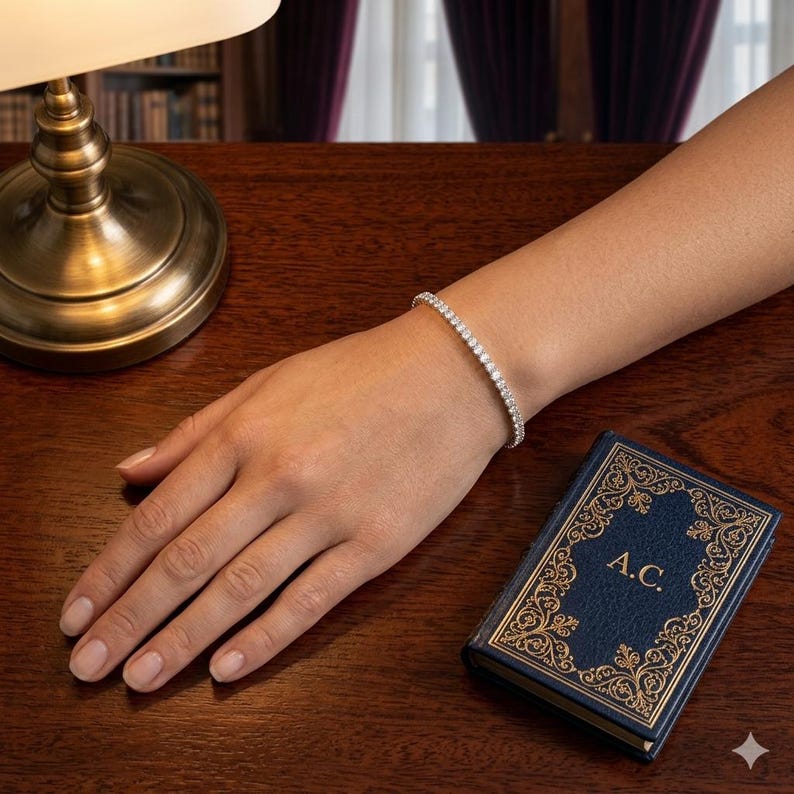 May include: A silver-toned tennis bracelet featuring a continuous row of small, clear, round-cut stones. The bracelet is worn on a wrist, positioned near a small, rectangular, dark blue book with gold-colored ornate borders and the letters A.C.
