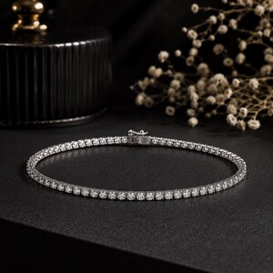 May include: A silver-toned tennis bracelet featuring a continuous row of round, clear, faceted stones set in a metallic frame. The bracelet includes a secure box clasp mechanism with a safety latch, showcasing a uniform design throughout the piece.