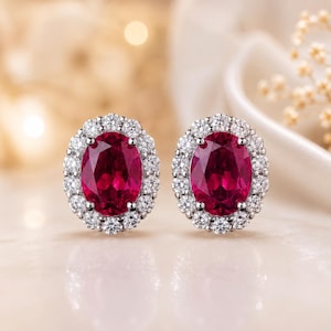 Può includere: Un paio di orecchini con rubini ovali circondati da un'aureola di piccoli diamanti rotondi. I rubini sono di un rosso intenso e i diamanti brillano. Gli orecchini sono incastonati in un metallo chiaro, forse platino o oro bianco.