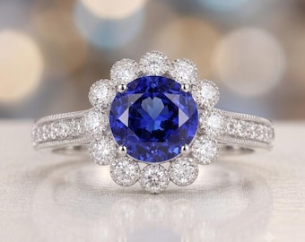 Puede incluir: Un anillo con una gran piedra central redonda de color azul intenso rodeada por un halo de doce piedras pequeñas, transparentes y de corte redondo. La banda es metálica con borde milgrain y tiene una hilera de piedras diminutas y claras.