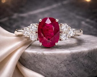 Op de afbeelding: Een ring met een grote, ovaal geslepen, dieprode robijn in het midden, vastgezet met vier pootjes. De ringband is gemaakt van gepolijst zilverkleurig metaal en is aan weerszijden versierd met een cluster van drie kleine, ronde witte diamanten.