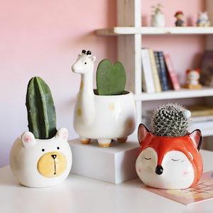 May include: Three ceramic animal-shaped planters: a bear, a giraffe, and a fox. The bear planter is white with a yellow face and a cactus. The giraffe planter is white with yellow spots and a green plant. The fox planter is red with a cactus.