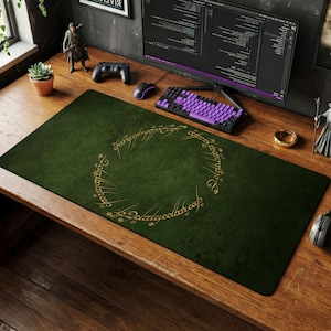 May include: A rectangular dark green desk mat featuring a circular gold-colored inscription in the center. The mat has a textured surface and is positioned on a wooden desk surface, serving as a base for a computer keyboard and mouse setup.