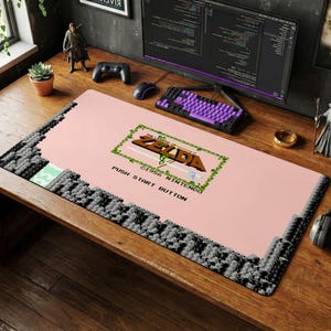 May include: A rectangular desk mat featuring a light pink background with a pixelated gray stone border. The center displays the 1986 Nintendo The Legend of Zelda logo framed by green vines, with the black text PUSH START BUTTON printed below it.