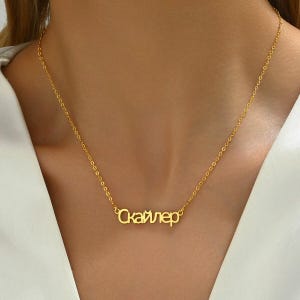 May include: A gold-colored necklace with the name "Скайлер" in a cursive font. The pendant hangs from a delicate chain. The necklace is displayed against a neutral background.