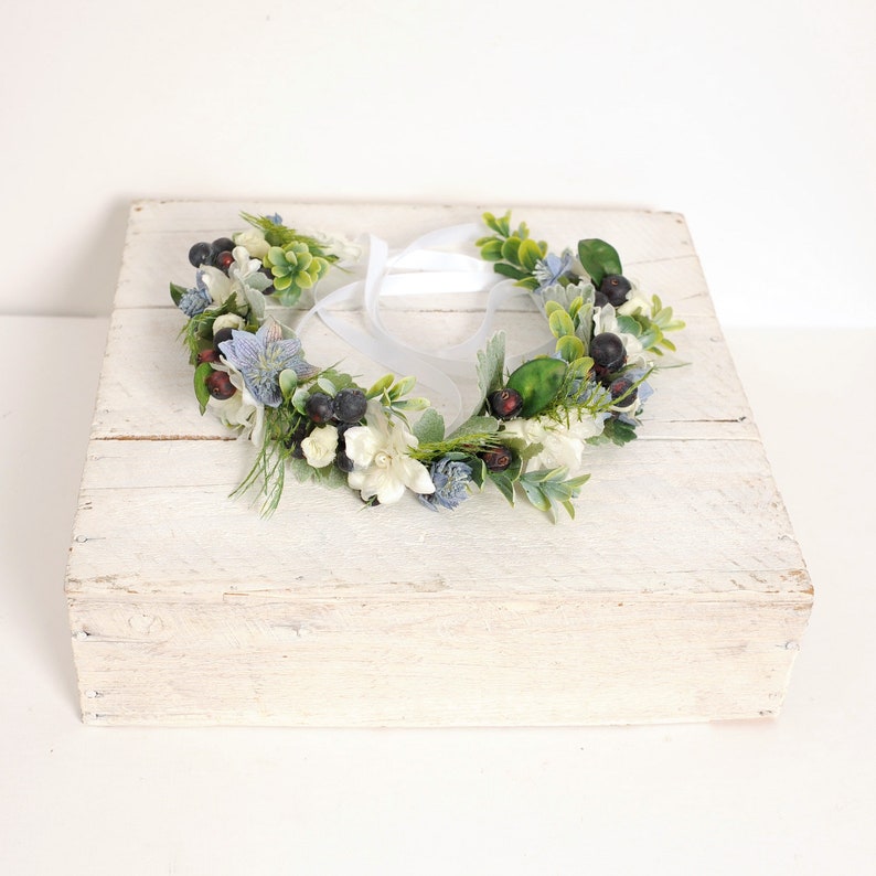 The photo shows a woodland style flower crown made with soft blue florals, green leaves and ferns, dark blue grape like berries and some added white filler flowers. Ribbon ties at the back make the crown fully adjustable to fit brides or bridesmaids.