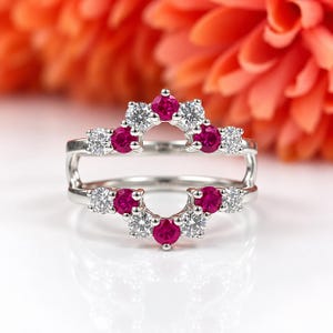 May include: A silver-toned ring guard featuring a double-band design with an open center. The piece is set with alternating round-cut clear white stones and vibrant pinkish-red stones arranged in a symmetrical, chevron-like pattern.