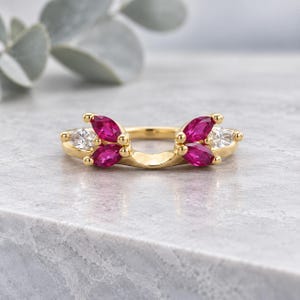 May include: A gold ring featuring a curved band design adorned with four marquise-cut vibrant pink gemstones and two smaller marquise-cut clear white gemstones, arranged symmetrically along the top of the polished yellow gold metal band.