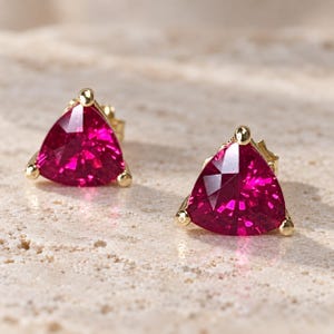 May include: A pair of stud earrings featuring triangular-cut, vibrant fuchsia-pink gemstones set in polished yellow gold. Each earring is secured with a three-prong setting, highlighting the faceted surface and deep colour of the stones.