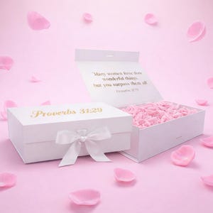 Puede incluir: Una caja de regalo blanca con el texto dorado "Proverbios 31:29", atada con una cinta blanca. La caja está abierta, revelando rosas rosas en su interior. El fondo es rosa con pétalos rosas esparcidos.