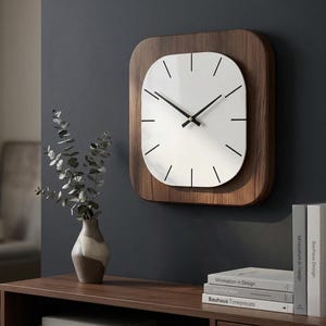 Puede incluir: Un reloj de pared moderno con esfera blanca y manecillas y marcadores negros. El reloj está enmarcado en un marco de madera rectangular. Está montado en una pared gris oscuro. Un jarrón con vegetación y libros están sobre una superficie de madera.