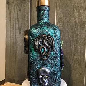 Könnte beinhalten: Eine dekorative Flasche mit einer strukturierten türkis-grünen Oberfläche, verziert mit einem goldfarbenen Verschluss. Die Flasche zeigt einen Drachen, einen Totenkopf und andere Verzierungen. Ein einzigartiger, handgefertigter Artikel.