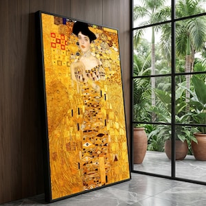 May include: Framed print of a Gustav Klimt painting. The artwork features a woman in a gold and yellow dress with intricate patterns. The frame is black, and the artwork is leaning against a wooden wall near a window with a tropical view.