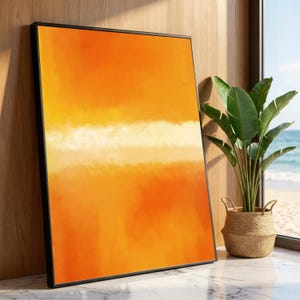 Puede incluir: Impresión artística abstracta en un marco negro. La obra presenta un degradado de tonos naranjas con una banda blanca horizontal. La pieza se exhibe en interiores, apoyada contra una pared de madera, con una planta en una cesta tejida cerca.