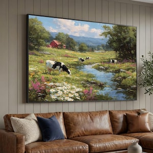 Könnte beinhalten: Ein gerahmtes Landschaftsgemälde zeigt eine ruhige ländliche Szene. Kühe grasen in einer grünen Wiese mit einem Bach, Wildblumen und einer roten Scheune unter blauem Himmel. Das Kunstwerk wird über einem braunen Ledersofa ausgestellt.