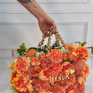 May include: A clear acrylic handbag filled with orange and peach-colored flowers. The bag has gold-colored chain handles and the name "ILKNUR" in gold lettering. A heart-shaped charm is also visible.