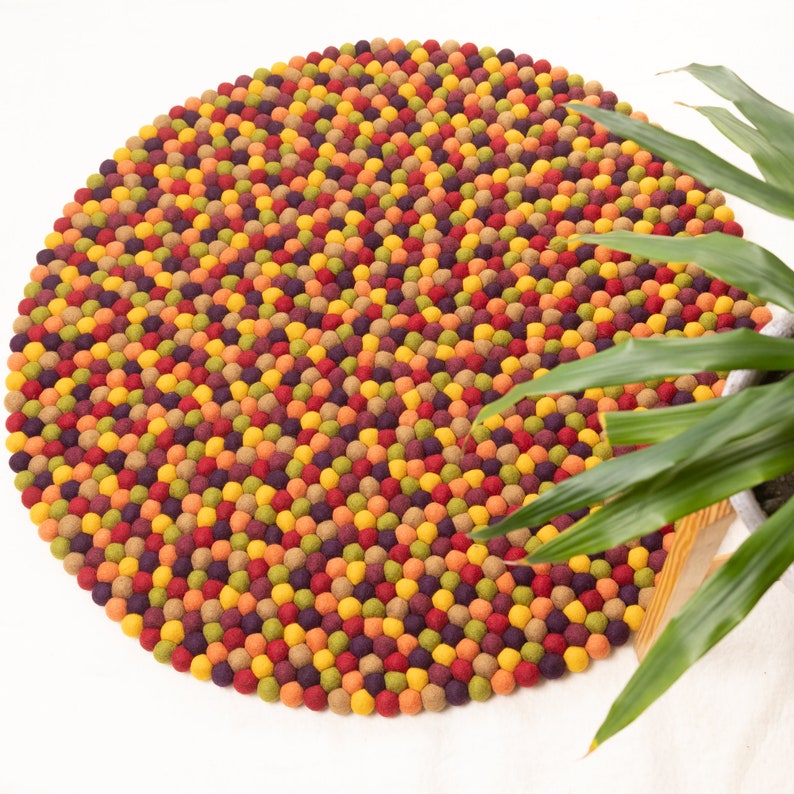 May include: A round, colorful felt rug made of many small balls. The rug is in shades of red, orange, yellow, green, and purple. The rug is on a white surface.