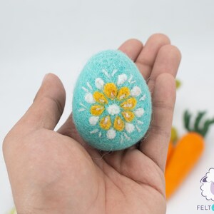 K&ouml;nnte beinhalten: Ein blaues Filz-Osterei mit einem wei&szlig;en und gelben Blumendesign. Das Ei wird in einer Hand gehalten.