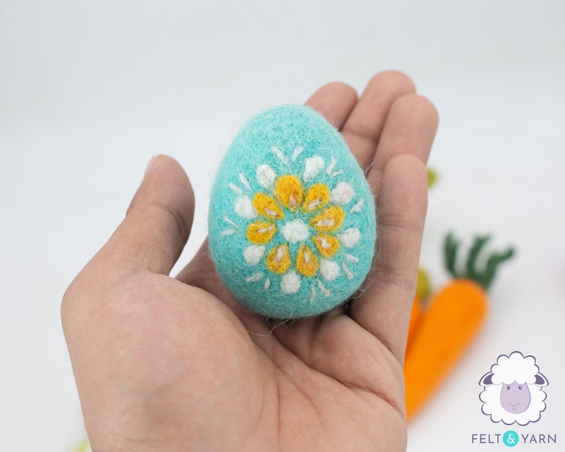 K&ouml;nnte beinhalten: Ein blaues Filz-Osterei mit einem wei&szlig;en und gelben Blumendesign. Das Ei wird in einer Hand gehalten.