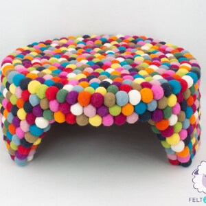 Pu&ograve; includere: Un pouf rotondo colorato fatto di palline di feltro. Il pouf ha un diametro di circa 45 cm e presenta una variet&agrave; di colori, tra cui rosso, arancione, giallo, verde, blu, viola e bianco.