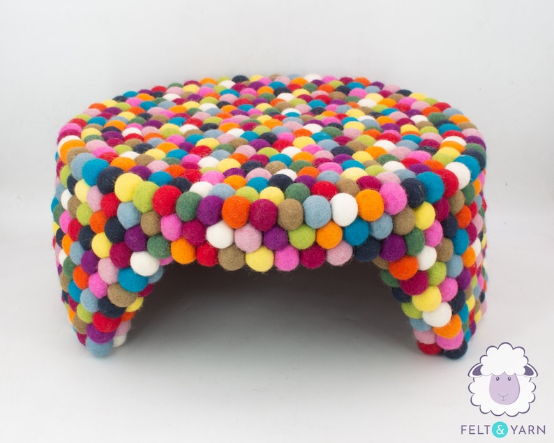 Pu&ograve; includere: Un pouf rotondo colorato fatto di palline di feltro. Il pouf ha un diametro di circa 45 cm e presenta una variet&agrave; di colori, tra cui rosso, arancione, giallo, verde, blu, viola e bianco.