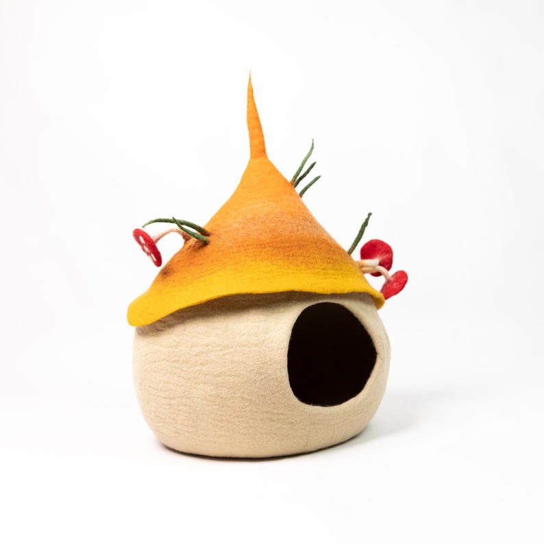 May include: A felted wool pet cave featuring a cream-colored rounded base with a large circular opening. The top is a pointed, cone-shaped roof that transitions from yellow to orange, decorated with small red mushroom accents and green stems.