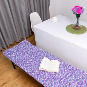 Pode incluir: Um assento de banco de pompons roxo, azul e branco com um livro em cima. O assento é feito de lã e tem uma superfície macia e texturizada.