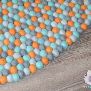 May include: A colorful felt ball rug with a pattern of orange, teal, gray, and white spheres. The rug is made of soft, round felt balls and is suitable for a nursery or playroom. The rug is on a wooden surface.