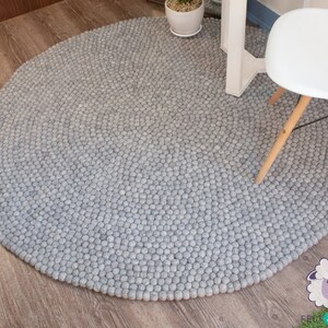 K&ouml;nnte beinhalten: Ein runder grauer Filzteppich aus kleinen Wollkugeln. Der Teppich liegt auf einem Holzboden und ein wei&szlig;er Stuhl mit Holzbeinen steht darauf.