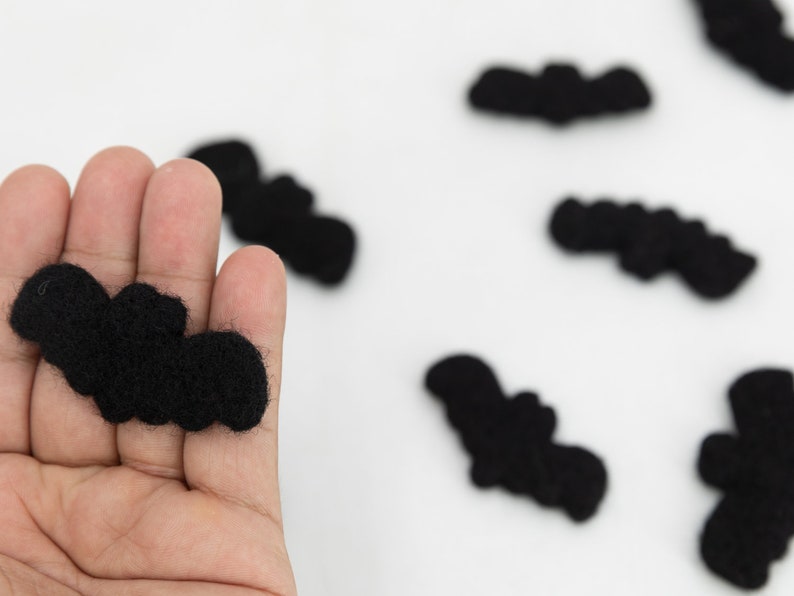 May include: A hand holding a black felt bat, with several other black felt bats scattered on a white surface.