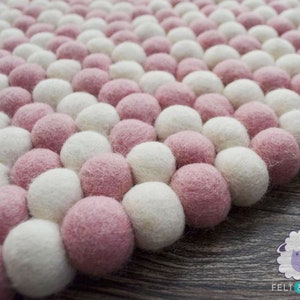 May include: A close-up of a pink and white pom-pom rug. The rug is made of soft, fluffy balls of wool felt.