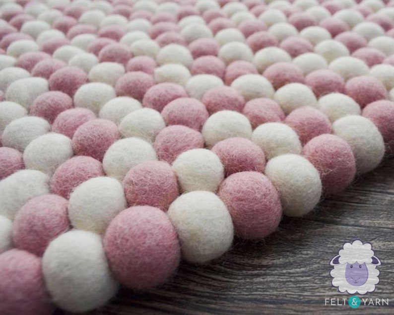 May include: A close-up of a pink and white pom-pom rug. The rug is made of soft, fluffy balls of wool felt.