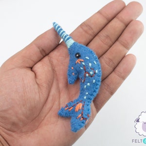 May include: A blue felt narwhal with a white and blue striped horn, orange and white embroidered details, and black embroidered eyes.