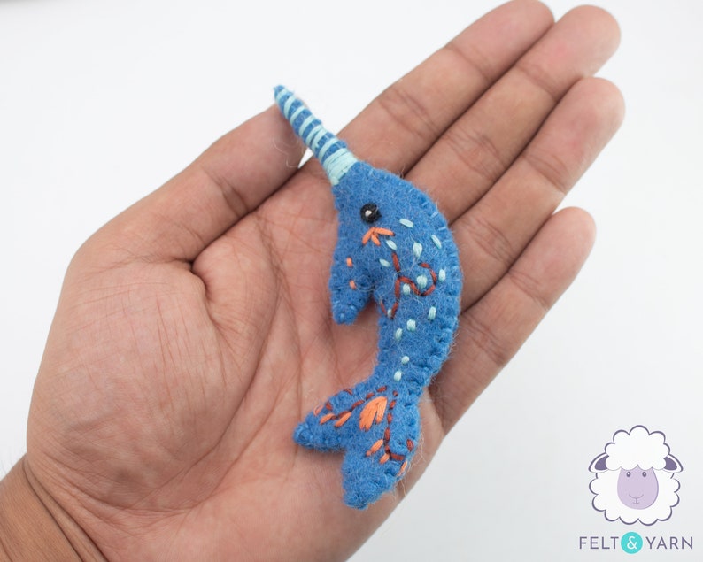 May include: A blue felt narwhal with a white and blue striped horn, orange and white embroidered details, and black embroidered eyes.