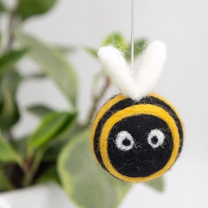 Op de afbeelding: Een vilt bijen ornament met zwarte en gele strepen, witte vleugels en zwarte ogen. De bij hangt aan een wit touwtje.