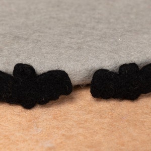 May include: Two black felt bat shapes on a light brown surface. The bats are about 2 inches long and 1 inch wide.