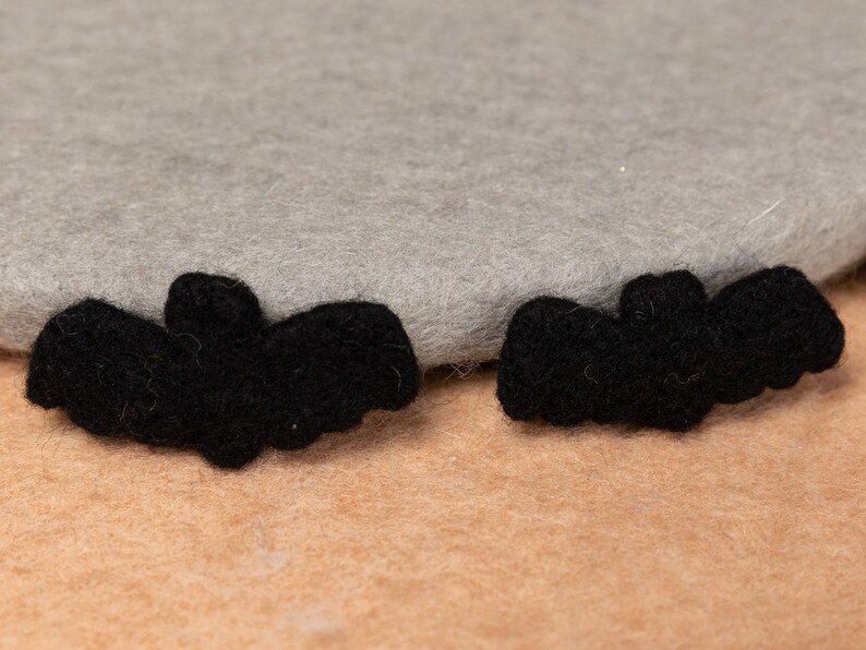 May include: Two black felt bat shapes on a light brown surface. The bats are about 2 inches long and 1 inch wide.
