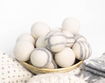 May include: A collection of round felted wool balls in a small woven basket. The balls are primarily solid off-white, with several featuring thin, horizontal gray stripes. The texture of the wool is soft and matte, creating a uniform appearance.