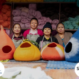 May include: Four women stand in front of a wall of colorful yarn, holding handmade felt cat beds in orange, blue, and red. The beds are shaped like teardrops and have a hole for the cat to enter. The women are wearing aprons and smiling. The text "goodweave+ CERTIFIED" and "Sedex AUDITED" is visible in the image.