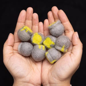 May include: A collection of gray felt balls with yellow accents. The balls are arranged in a pile and are being held in the palm of a person's hand.