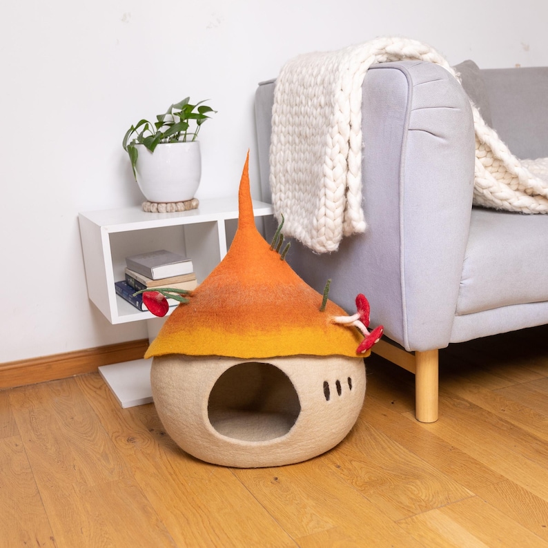 May include: A felted pet cave shaped like a mushroom with a beige base and a gradient orange and yellow cap. The cap features small decorative red mushroom accents and green stems. The structure has a circular entrance and three vertical slits.