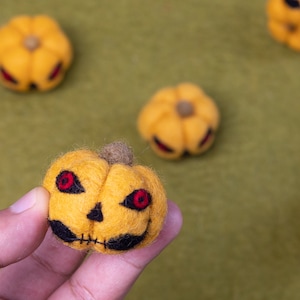 May include: A close-up of a hand holding a small, yellow felt pumpkin with a stitched-on face and red eyes. The pumpkin is surrounded by other felt pumpkins on a green surface.