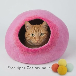 May include: A bright pink felted wool cat cave with a round opening containing an orange tabby cat. Next to the cave are four small felted wool balls in yellow, orange, and light green colors. The text reads: Free 4pcs Cat toy balls.