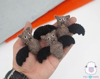 May include: Three brown felt bats with black felt wings. The bats have white felt eyes and are being held in a hand.